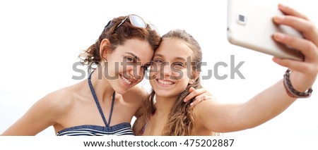 Panoramic portrait of young sisters on a summer beach holiday joyfully smiling with heads together taking selfie photo, holding up a smart phone, outdoors. Travel technology lifestyle.