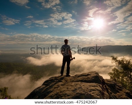 Tall photographer with baseball cap and tripod with camera in hands stand on rocky view point and watching down to deep misty valley bellow