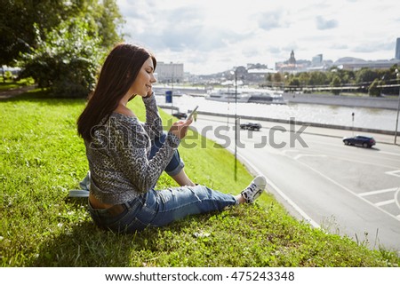 Attractive young white brunette girl sitting on green grass with city view using her modern smartphone with touch screen. Always stay connected to internet with your mobile phone 