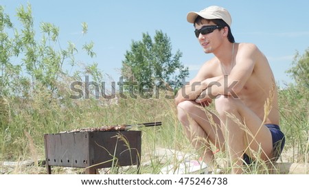 Young man in sunglasses fry the meat on the beach near the sea. Hot food Shashlik in Russia.