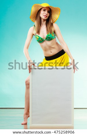 Holidays summer and advertisement concept. Woman wearing yellow hat and bikini holding blank presentation board. Female model posing in full length on blue background.