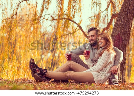 Young couple in love sitting on a fallen autumn leaves under a tree in a park, drinking an ice tea and enjoying a wonderful autumn day