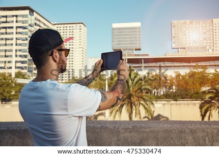 Back shot of a young man in plain white t-shirt and baseball hat taking a photo of city buildings and palm trees on his tablet.