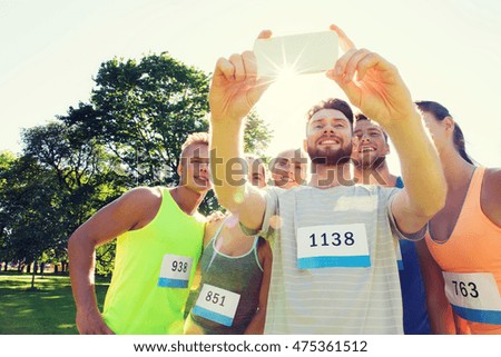 fitness, sport, friendship, technology and healthy lifestyle concept - group of happy sportsmen friends with racing badge numbers taking selfie smartphone outdoors