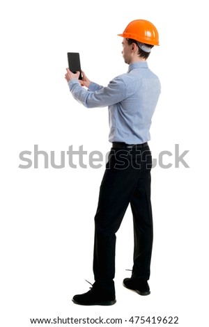 Backview of business man in construction helmet stands and enjoys tablet or using a mobile phone. Curly businessman in a light jacket and a helmet makes selfie tablet.