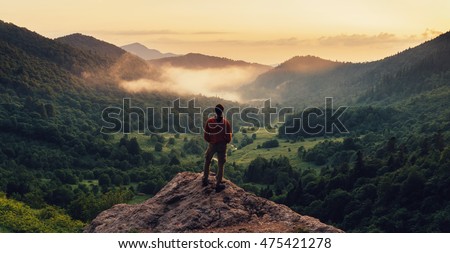Young man standing on top of cliff in summer mountains at sunset and enjoying view of nature Royalty-Free Stock Photo #475421278