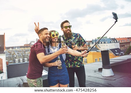Bearded friend giving rabbit ears to man with beer while woman in sunglasses holds a camera phone at the end of a selfie stick