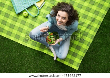 Smiling woman having a relaxing lunch break outdoors, she is sitting on the grass and eating a salad bowl Royalty-Free Stock Photo #475492177
