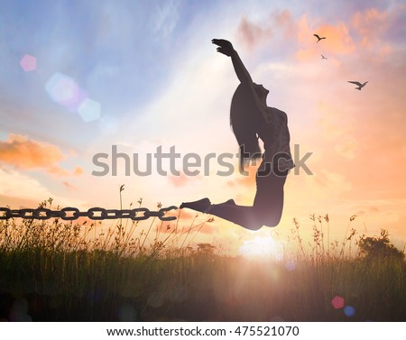 International day for the remembrance of the slave trade and its abolition concept: Silhouette of a woman jumping and broken chains at orange meadow autumn sunset with her hands raised Royalty-Free Stock Photo #475521070