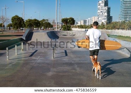 Backshot of a longboarder in white plain t-shirt walking with his longboard in a concrete skate park against the background or city buildings, his dog following him