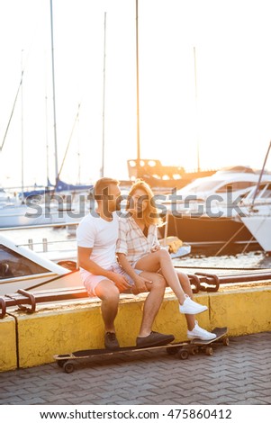Young beautiful couple sitting at seaside, smiling, skateboarding.