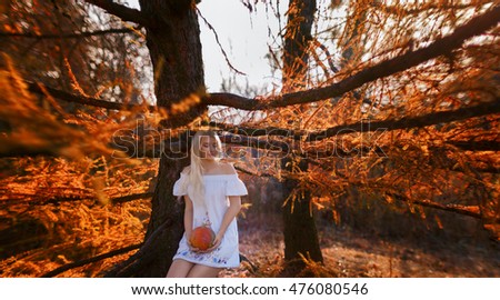 A woman dressed in a white dress with embroidery holds a pumpkin hands. She sits on the branch of spruce. Sunlight painted the picture in warm tones.