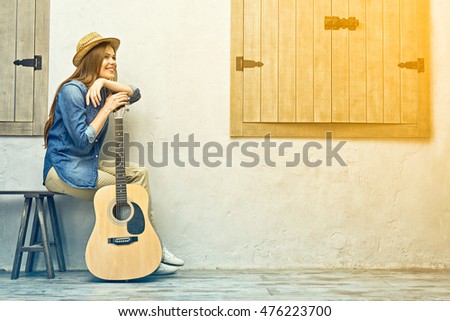 Smiling woman with guitar sitting on street bench against wall with windows.