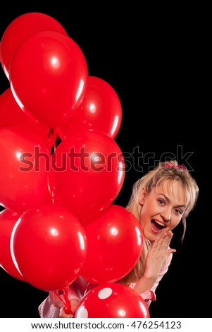 Happy young blond woman looking out of red balloons and smiling with admiration. Isolated on black background