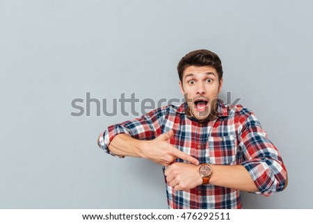 Amazed young man in plaid shirt pointing on wristwatch over grey background Royalty-Free Stock Photo #476292511