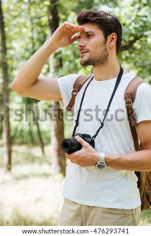 Handsome young man photographer holding modern photo camera and looking far away in forest