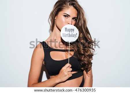 Image of a young brunette girl in bikini covering mouth with happy birthday sign on stick over white background