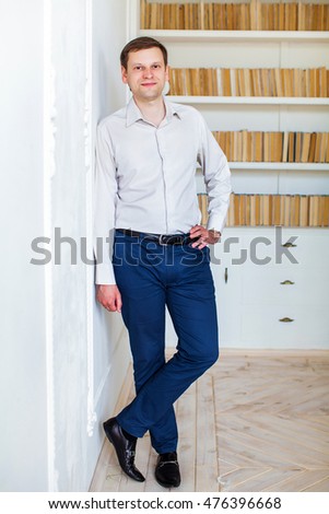 Stylish business man in white shirt and trousers, standing against the wall, against the background of shelves of books