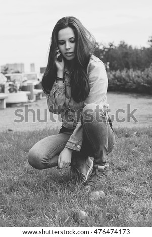 Beautiful a young girl in a denim jacket sitting in the park and talking on her smart phone. Photo in black and white style. 