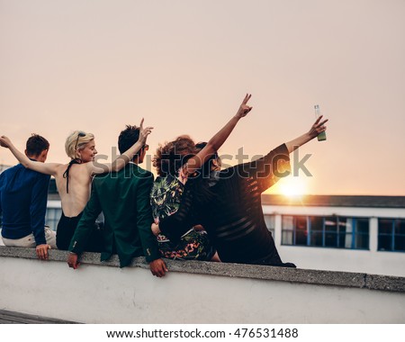 Rear view of young people partying on terrace with drinks at sunset. Young men and women enjoying drinks on rooftop in evening. Royalty-Free Stock Photo #476531488