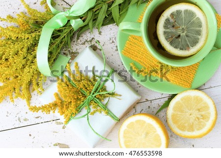Fresh composition with cup of lemon tea, gift box wrapped with rough white paper and decorated with jute and bunch of bright yellow flowers on wooden table. Romantic dreams
