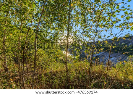 sunset at quarry or lake or pond with sandy beach, green water, birch on a foreground and hills with blue sky at summer season