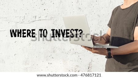 Young man using laptop and WHERE TO INVEST? concept on wall background