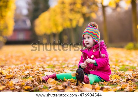 Happy little girl in pink warm coat and colorful knitted hat playing in beautiful autumn park on warm sunny fall day. Kids play with golden maple leaves.
