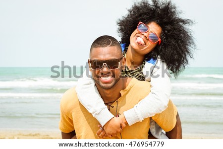 Young afro american couple playing piggyback ride on beach - Cheerful african friends having fun at day with blue ocean background - Concept of lovers happy moments on summer holiday - Vintage filter Royalty-Free Stock Photo #476954779