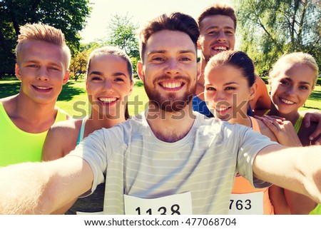 fitness, sport, friendship, technology and healthy lifestyle concept - group of happy teenage friends with racing badge numbers taking selfie by smartphone at marathon outdoors