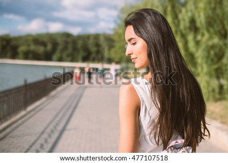 Close-up portrait of young brunette woman with long hair, with backpack, looking down and dreaming. Travel. Park background.