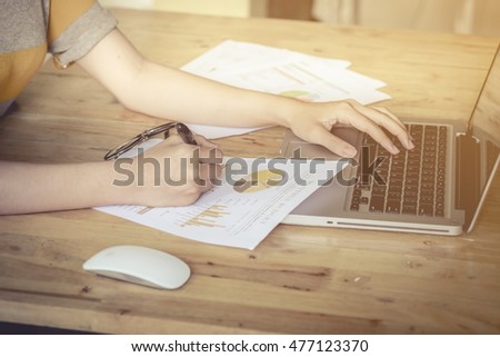 Business girl hand holding pen point stock market chart and using laptop on wood desk. vintage effect.