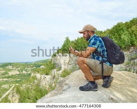 Man tourist is using a smartphone while sitting on the edge of a cliff in the mountains. Caucasian male outdoors in nature. Hiker young man with backpack .