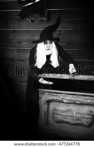 Old man wizard with long grey hair beard in black costume and hat for Halloween standing near wooden chest or trunk box on log house background, black and white