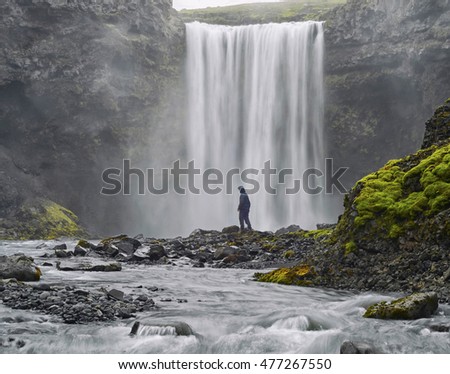 A man dressed in black standing at the base of a waterfall . Beautiful vibrant panorama picture with a view on icelandic waterfall