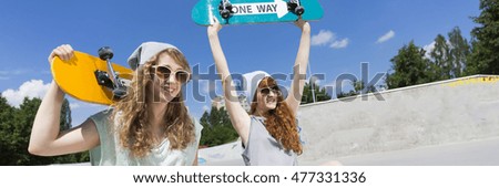 Two young female friends holding their skateboards, having fun in the park, panorama