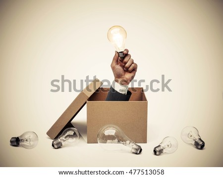 Hand of a businessman holding a turned on light bulb coming out from a brown paper box surrounded by old incandescent light bulbs / Business with new idea and innovation concept