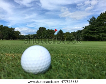 A white golf ball sits in the foreground just off the green of a course on a beautiful autumn day. Chip or putt hit into the cup.