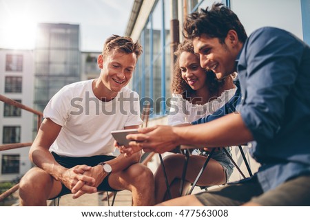 Group of people watching video on the mobile phone while sitting at outdoor cafe. Three young friends sitting outdoors and looking at smartphone. Royalty-Free Stock Photo #477563008
