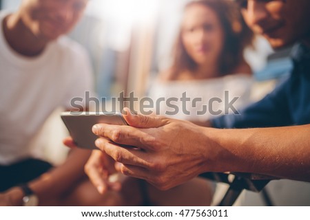 Young friends sitting outdoors and looking at smartphone. Young man showing something interesting to friends. Focus on mobile phone in hand. Royalty-Free Stock Photo #477563011
