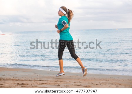 Young fit cute sporty girl in sportswear running on beach at sunset in morning. Fitness, workout and healthy lifestyle concept. Blue sky with clouds and sea on background