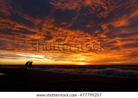 Sunset and dramatic clouds over the sea casts a glow on some indistinct human figures.