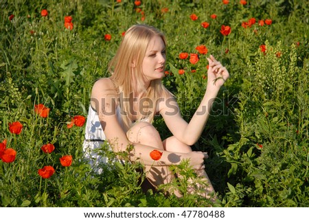 The girl sits on in the poppies field and holds a poppy in a hand