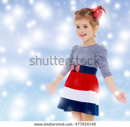 Little Caucasian blond girl with a red bow on her head, dressed in striped dress, she shrugs. Close-up.Gentle blue Christmas background with white snowflakes abstract.