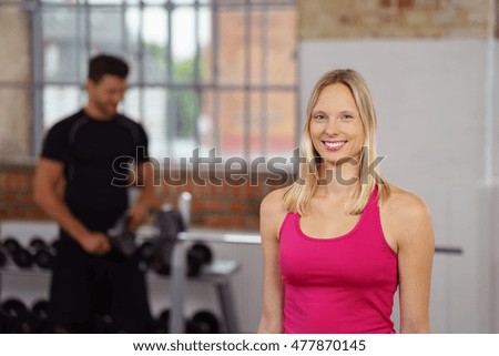 Cute smiling woman dressed in pink sleeveless top standing near fitness trainer in front of barbell on rack in gym with copy space
