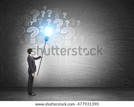 Businessman in suit looking at blackboard with question marks on it holding blue light bulb on stick. Concept of solution finding for challenging problem
