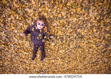 Little girl lying on a carpet of fallen autumn leaves in black jacket and sunglasses photoed from the top down