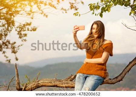 Cute happy young blonde Caucasian woman in denim jeans and brown sweater, taking a selfie outdoors in park on windy autumn day. Retouched, natural light.