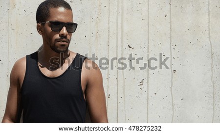 People and lifestyle concept. Portrait of good-looking African athlete with muscular arms standing by concrete wall, getting ready for his evening run, making up his mind before hard workout
