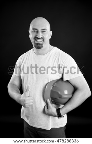 Black & white picture of a happy handsome man showing thumb up sign. Hardhat under his arm. Happy bald foreman with a goatee beard, isolated.
